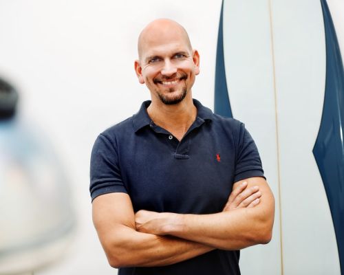 Zahnarzt Philipp Heitmüller steht lächelnd in seiner Praxis vor einem Surfbrett