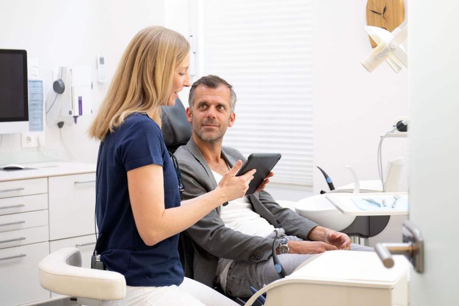 Zahnärztin Dr. Heinkele im Gespräch mit einem Patienten in der Praxis Heitmüller in Hannover. Sie hält ein Tablet, um den Befund zu zeigen.
