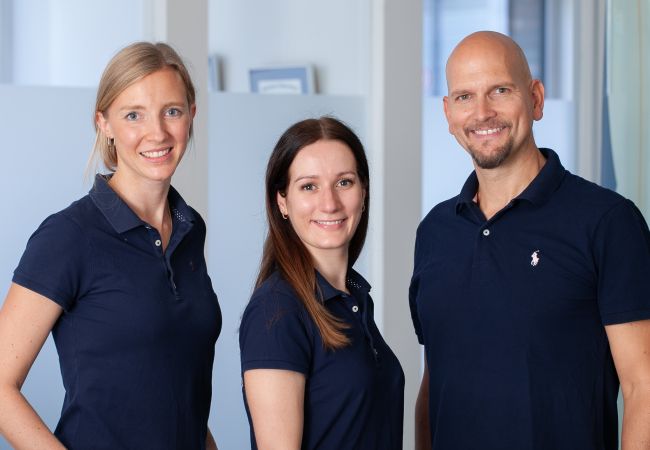 Drei Zahnärzte der Zahnarztpraxis Heitmüller, Hannover List, in dunkelblauer Teamkleidung vor einer Glaswand im Empfangsbereich.