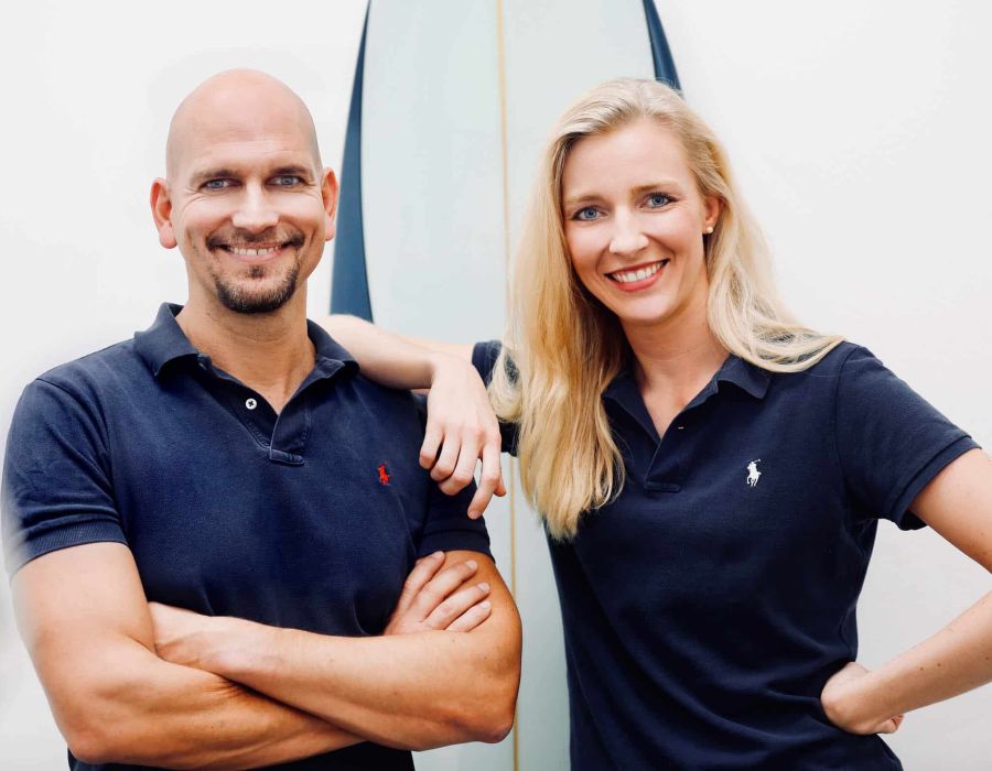Zahnarzt Philipp Heitmüller und Zahnärztin Dr. Laura Heinkele stehen gemeinsam vor einem Surfbrett in der Praxis
