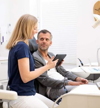 Zahnärztin Dr. Heinkele im Gespräch mit einem Patienten in der Praxis Heitmüller in Hannover. Sie hält ein Tablet, um den Befund zu zeigen.