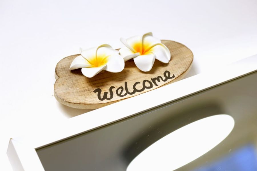 Holzschild mit der Aufschrift Welcome. Als Verzierung dienen zwei weiß-gelbe Frangipani-Blüten.