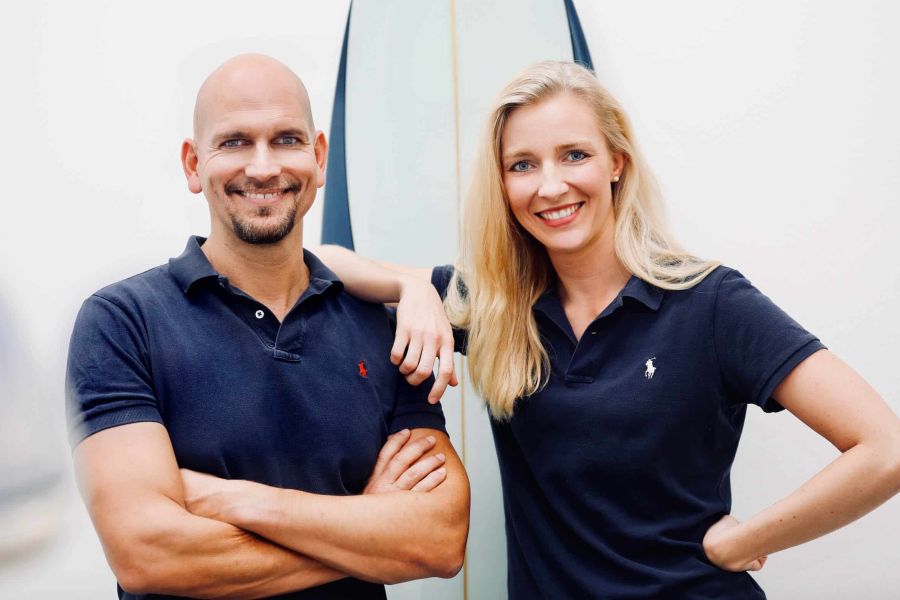 Zahnarzt Philipp Heitmüller und Zahnärztin Dr. Laura Heinkele stehen gemeinsam vor einem Surfbrett in der Praxis