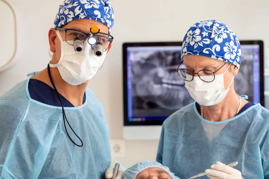 Hochkonzentrierte Teamarbeit bei einer Operation in der Zahnarztpraxis Heitmüller. Zahnarzt Philipp Heitmüller mit Lupenbrille und eine Assistentin blicken auf einen Patienten. Im Hintergrund sieht man das Röntgenbild.